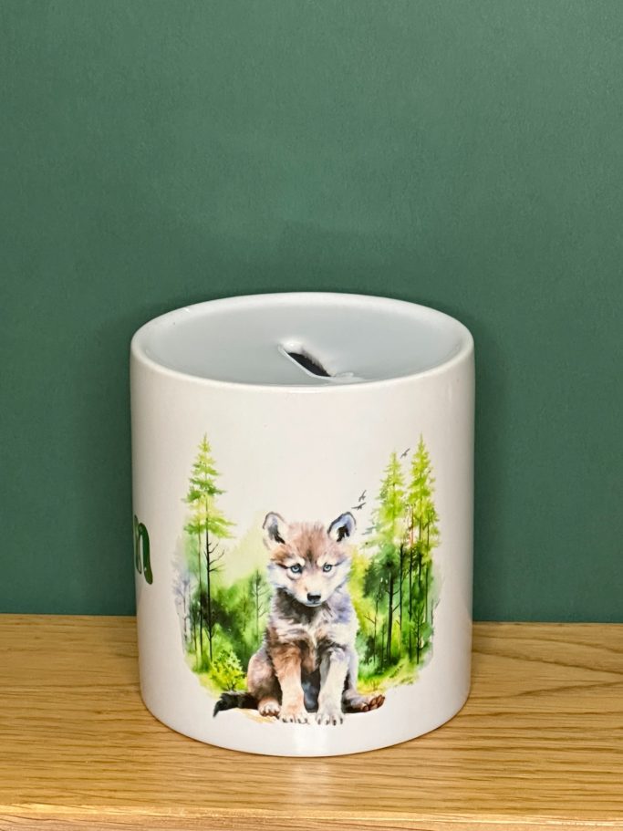 Spardose aus Keramik mit Wolf vor Wald Motiv. Schlitz in Oberseite, in Unterseite Loch mit Kunststoffdeckel zum Verschließen der Spardose.